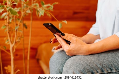 Side View Of Female Hands Holding Black Mobile Phone, Indoors. Close-up Of Caucasian Woman Chatting, Typing, Surfing Internet On Smartphone While Sitting On Couch At Home. Selective Soft Focus.