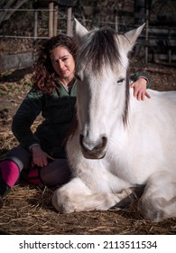 Side View Of Female Horse Trainer  In Green Polar Fleece Jacket And Pink Boots And Her White Pony Laying Down.