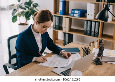 Side View Of Female Lawyer Doing Paperwork At Workplace With Laptop In Office