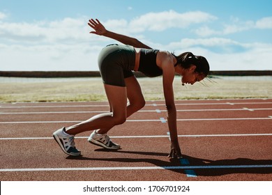 Side View Of Female Runner Getting Ready At The Start Line On Running Track At Stadium. Woman Athlete Taking Position On Her Marks To Start Off The Run.