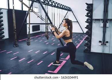 Side View Of Female In Sportswear Sitting Squat On One Leg While Holding Trx Fitness Straps At Stylish Empty Gym