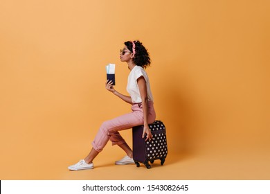 Side View Of Female Tourist Sitting On Suitcase. Studio Shot Of Curly Woman In Pink Pants Holding Tickets On Yellow Background.