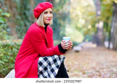 Side View Of Female Wearing Red Warm Outerwear Sitting With Cup Of Takeaway Hot Drink In Autumn Park And Looking Away