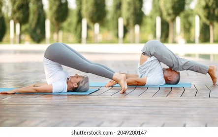 Side view of fit active senior family couple in sportswear doing plow pose during yoga training in park on summer day, mature man and woman practicing Halasana on mat outdoors
