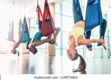 Side view of a fit and beautiful young woman hanging upside down while practicing aerial yoga during group class in a modern fitness club