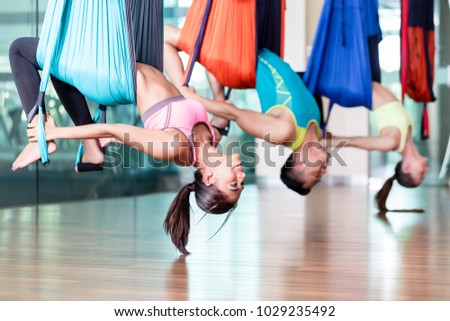 Side view of a fit and beautiful young woman hanging upside down while practicing aerial yoga during group class in a modern fitness club