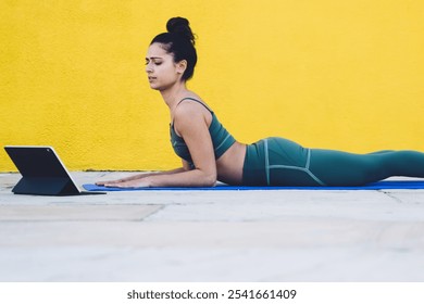 Side view of fit brunette in green sportswear outfit lying in low cobra pose on yoga mat and watching tutorial on tablet screen against yellow background - Powered by Shutterstock