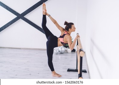 Side view of fit focused ballerina standing at barre holding onto handrail in bright studio and stretching body lifting leg in air 