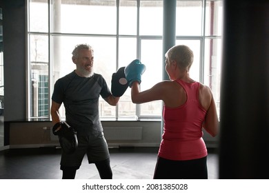 Side View Fit Middle-aged Woman In Pink Top Boxing With Personal Trainer In Punch Mitts. Experienced Fitness Trainer Energizes Woman To Workout Harder. Stress Relief Activities.