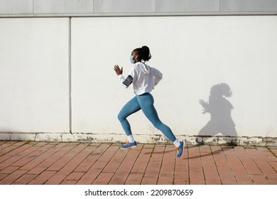 Side View Of Fit Sporty Woman Running On Asphalt Wearing Face Mask Against Coronavirus Covid-19. Female Athlete Training Outside Under Pandemic.