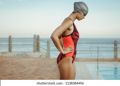 Side View Of Fit Woman Swimmer Concentrating At Pool With Her Hands On Hips. Competitive Female Swimmer Wearing Swim Cap And Goggles Near Swimming Pool At Seaside.