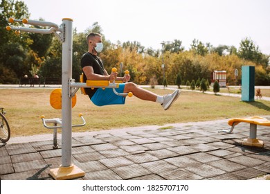 Side View Of Fit Young Male Caucasian Athlete With Face Mask Trains On Arm And Leg Equipment In An Outdoor Gym. Muscular Athlete Outdoors.  COVID - 19 Coronavirus Protection