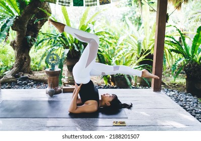 Side View Of Flexible Asian Girl Practicing Yoga On Wooden Terrace. Concept Of Healthy Lifestyle. Young Concentrated Woman Wearing Sportswear And Barefoot On Fitness Mat. Bali Island. Sunny Day