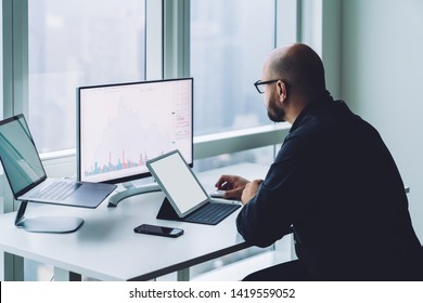 Side View Of Focused Bearded Man In Glasses Sitting With Tablet And Laptop At Desktop While Working On Computer In Office