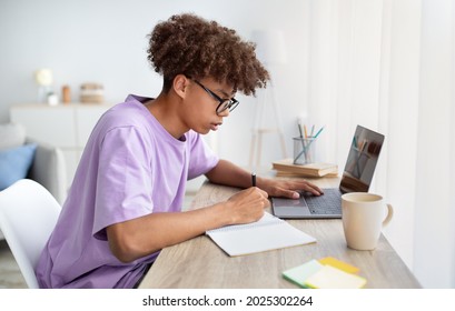 Side view of focused black teenager in glasses studying online from home, writing in notebook, using laptop pc at table indoors. E-learning, web-based education during covid lockdown