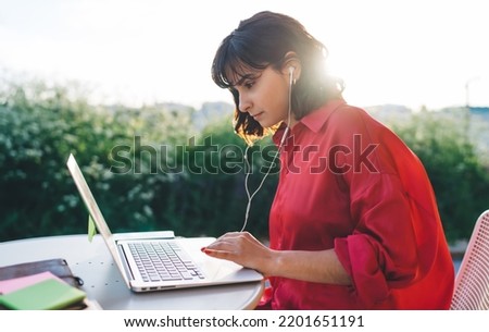 Side view of focused brunette with short hair wearing red shirt and earphones browsing laptop while working remotely at table in sunny garden