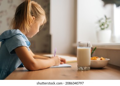 Side view of focused elementary little child girl learning sitting at home table with cookies and milk by window. Close-up of preschool student writing in copybook studying alone, selective focus.