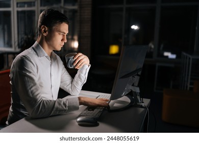 Side view of focused male reading on laptop working on deadline during late shift. Business, online and corporate worker drinking coffee and doing research on computer during overtime at work. - Powered by Shutterstock