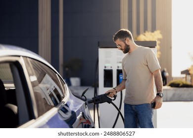 Side view of a focused man plugging a charger into his electric car at a charging station, with modern city architecture in the background.