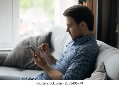 Side View Focused Man Using Smartphone, Sitting On Couch At Home Alone, Young Male Looking At Phone Screen, Chatting In Social Network With Friends, Shopping Online, Browsing Mobile Device Apps