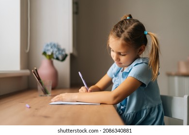 Side view of focused primary little child girl learning writing doing homework sitting at home table by window. Portrait of smart preschool kid studying alone making note with pen at bedroom.