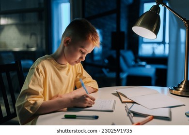 Side view of focused schoolboy studying at home doing homework sitting at table under light of lamp. Smart redhead pupil boy writing in exercise book at night in dark with lighting lamp.