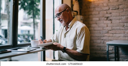 Side View Of Focused Senior Bearded Male Journalist In Casual Clothes Working Near Window In Modern Cafe And Taking Notes In Notepad