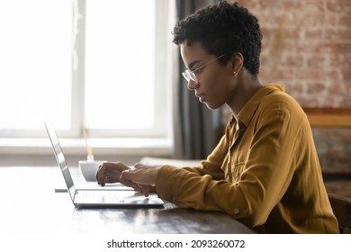 Side view focused young African American businesswoman in eyewear working on computer, sitting at table in modern loft office room, typing message, preparing electronic document or presentation.
