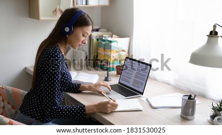 Side view focused young beautiful caucasian woman wearing wireless headphones, studying remotely using laptop at home office, smart millennial female student involved in research article writing.