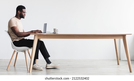 Side view of focused young black man using laptop, sitting at desk, working or studying online from home office, blank space. Full length of African American man having remote freelance job