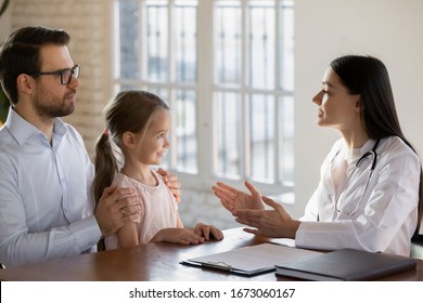 Side View Focused Young Father Holding On Lap Smiling Little Daughter Listening To Female General Practitioner At Checkup. Professional Medical Worker Explaining Lab Test Results To Patients.