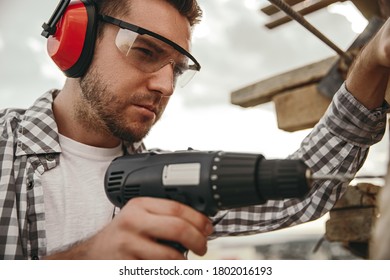 Side view of focused young male worker in protective goggles and headphones using electric drill while working on construction site