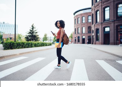 Side View Of Focused Youthful Attractive Black Woman Looking At Camera And Using Smartphone While Walking On Pedestrian Crossing In Town Waiting For Taxi After Order Via App