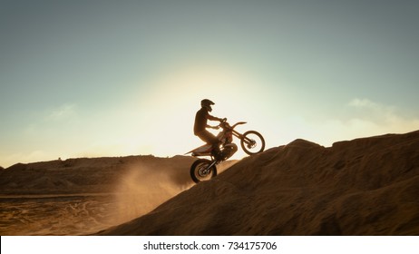 Side View Footage of the Professional Motocross Motorcycle Rider Driving on the Dune and Further Down the Off-Road Track. It's Sunset and Track is Covered with Smoke/ Mist.