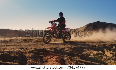 Side View Footage of the Professional Motocross Motorcycle Rider Driving on the Dune and Further Down the Off-Road Track. It's Sunset and Track is Covered with Smoke/ Dust/ Dirt.