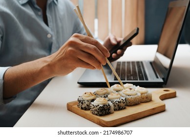 Side view of freelancer working on laptop, using smartphone in home office while sitting at table during lunch, close-up. Selective focus on male hand holding chopsticks and sushi. - Powered by Shutterstock