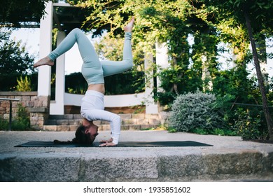Side view full body attractive young female in sportswear doing headstand exercise with splits on mat while practicing yoga in summer park