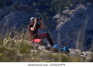 Side view full body of male hiker sitting on rocky cliff and looking away through binoculars against mountains on sunny day - Powered by Shutterstock