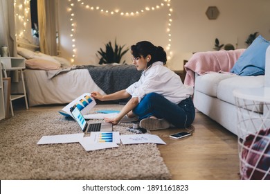 Side view full length attractive female wearing casual clothes doing paperwork and browsing modern netbook while sitting on floor with crossed legs in cozy bedroom