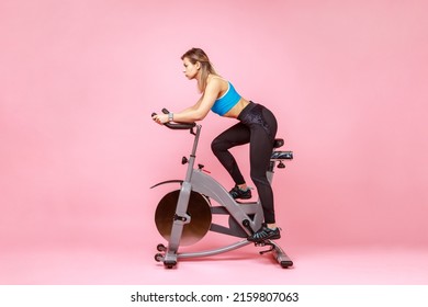 Side View Full Length Of Beautiful Sportswoman Cycling A Bike At Home, Cardio Training, Exercising Legs, Wearing Sports Tights And Top. Indoor Studio Shot Isolated On Pink Background.