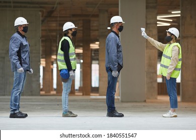 Side View Full Length Of Female Supervisor Measuring Temperature Of Workers With Contactless Thermometer At Construction Site, Corona Virus Safety Measures