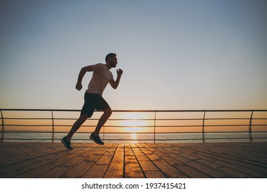 Side View Full Length Portrait Of Handsome Attractive Young Bearded Athletic Man Guy 20s In Casual White T-shirt Black Shorts Posing Training Running Looking Aside At Sunrise Over The Sea Outdoors