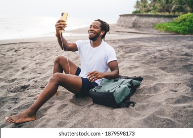 Side view full length smiling African American male in summer shorts resting on sandy tropical beach and having video call through mobile phone - Powered by Shutterstock