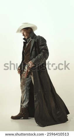 Side view full-length image of serious man, cowboy in leather trench coat and hat, standing with thoughtful gaze against light background. Concept of American culture, history, wild west