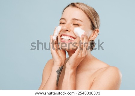 Side view fun beautiful half naked topless young woman with nude make up apply soap foam for washing face isolated on plain blue color background. Skin care spa healthcare cosmetic procedures concept