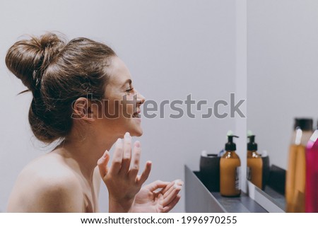 Side view fun half naked topless young woman 20s applying moisturizer, anti-aging facial cream stand in front of mirror in bathroom do morning routine Skin care healthcare cosmetic procedures concept