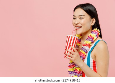 Side view fun happy young woman of Asian ethnicity in striped one-piece swimsuit hawaii lei drink soda pop cola fizzy water isolated on plain pastel pink background. Summer vacation sea rest concept