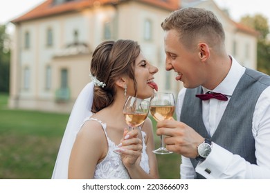 Side View Of Funny Wedding Couple Standing Outside Near Old Building In Park In Summer, Showing Tongues To Each Other. Young Man Groom And Woman Bride Raising Hands With Glasses Of Wine. Celebration.