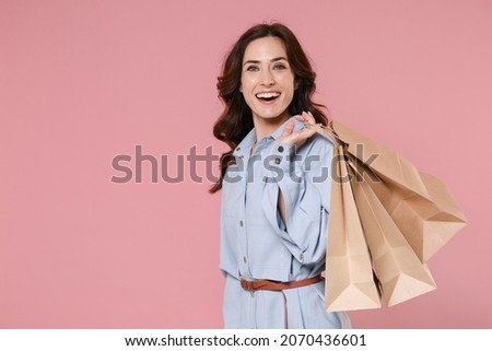 Side view of funny young brunette woman 20s wearing casual blue shirt dress hold package bag with purchases after shopping looking camera isolated on pastel pink colour background, studio portrait