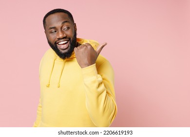 Side view of funny young man of African American ethnicity 20s wear casual basic yellow hoodie stand pointing thumb aside on mock up copy space isolated on pastel pink color background studio portrait
